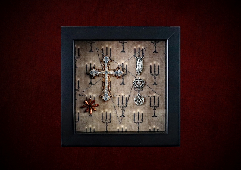 Repurposed black shadow box frame featuring a large diamante cross, Virgin Mary reliquary, and a pentagram charm on a candelabra backdrop.