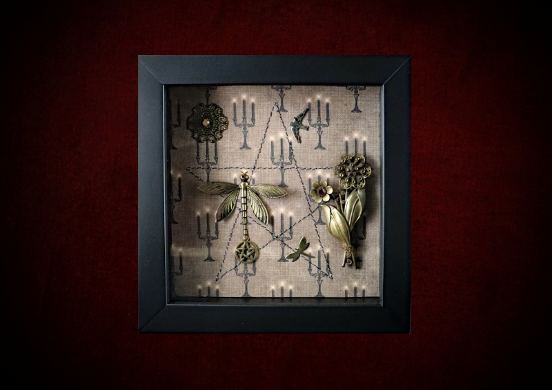 Repurposed black shadow box frame featuring a central dragonfly, flying bird charm, and a vintage mauve diamante brooch on a candelabra backdrop.