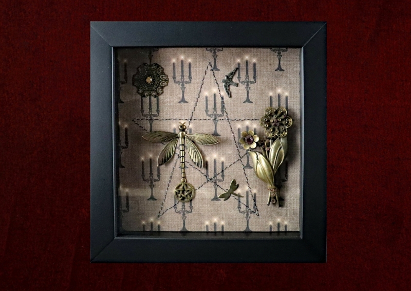 Detailed view of the central dragonfly charm flanked by smaller charms in flight inside a traditional gothic curiosity display frame.