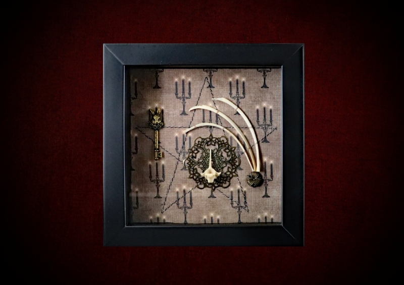 Repurposed black shadow box frame featuring hare rib bones, a hare's head key, and a gold filigree disc on a candelabra backdrop.